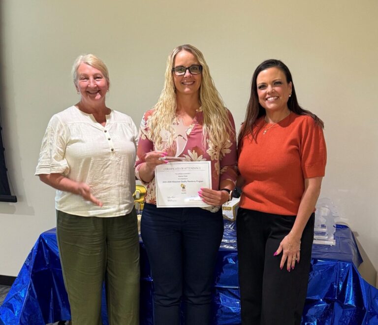 Three smiling women stand together indoors; the center woman holds a certificate of attendance while a blue wrapped table is in the background.