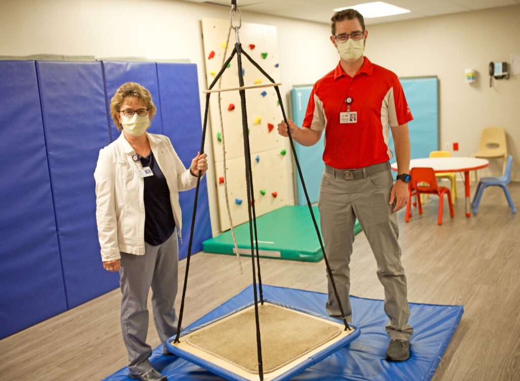 Two adults wearing masks stand beside a suspended provider harness in a rehab gym, blue mats visible behind them.