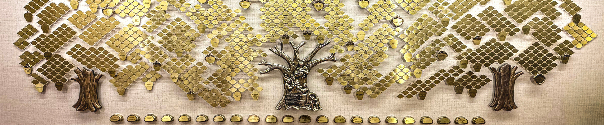 Gold display of diamond-shaped plaques forming trees on a beige fabric background, with a central tree sculpture and a row of small plaques at the base.