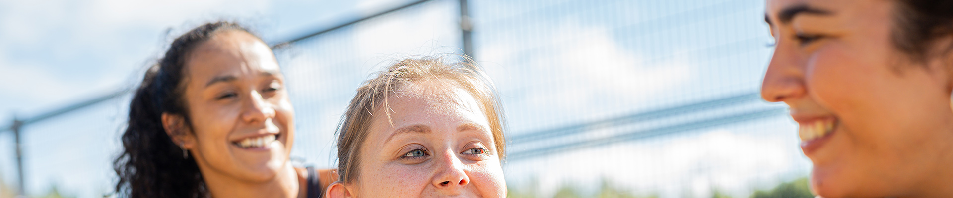 Three friends smiling and chatting outdoors on a sunny day, with a chain-link fence in the background.