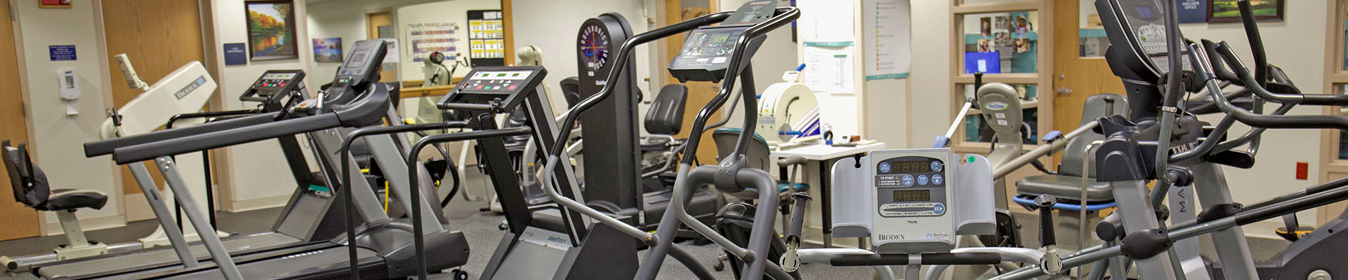 Cardio equipment in a gym: treadmills, ellipticals, and stationary bikes arranged in a fitness room with a medical or rehab setting backdrop, neutral lighting.