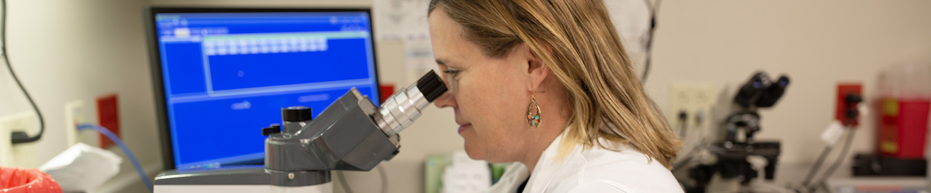 Female researcher looks into a microscope at a lab bench with a blue computer screen in the background