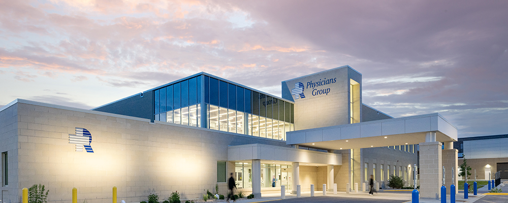 Modern medical clinic named Physicians Group at dusk, with glass atrium and a covered entrance.