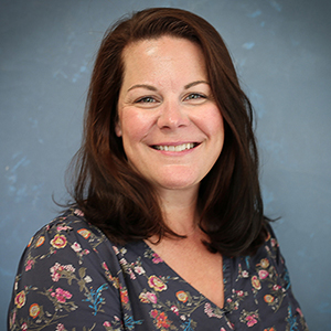 Portrait of a smiling woman with shoulder-length brown hair wearing a floral blouse against a blue textured background.
