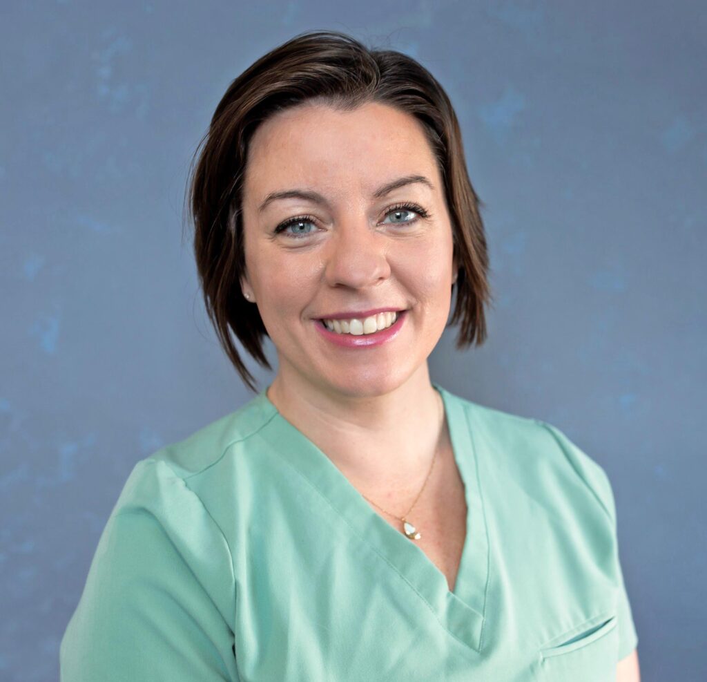 Smiling woman in green scrubs against a blue background, cropped to show the face and neckline.