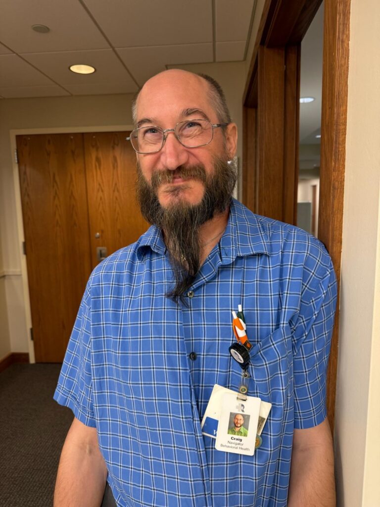 Bearded man with glasses in a blue plaid shirt wearing an employee ID badge, standing in an office hallway and smiling at the camera.