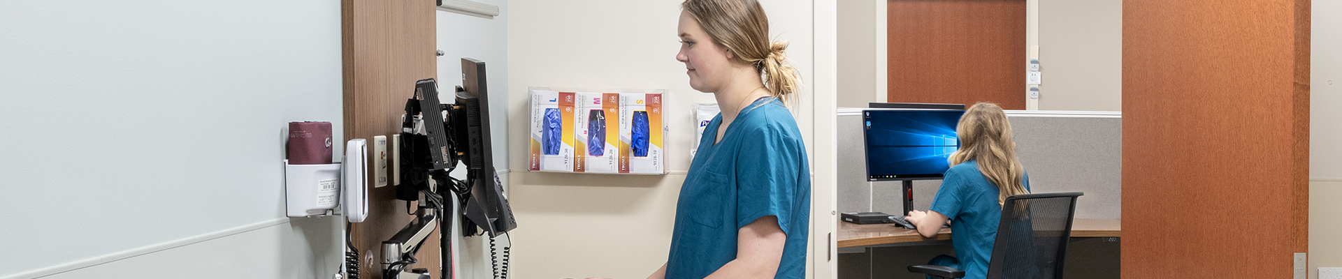 Nurse in blue scrubs standing at a clinic computer workstation with phones and monitors.