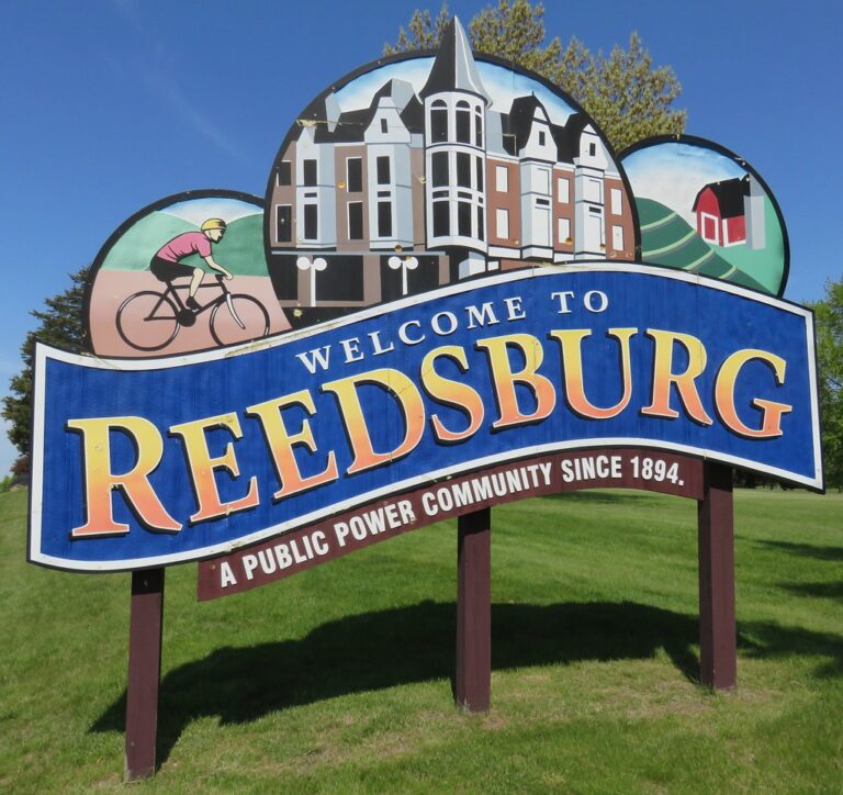 Welcoming sign for Redbsurg featuring a large 'REEDSBURG' text and a town illustration above, with 'Welcome to' and 'A Public Power Community since 1894' on a curved banner.