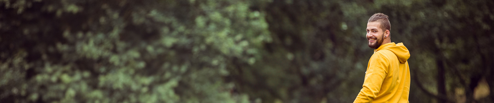 Man in a yellow hoodie smiling while walking through a wooded park setting, trees in the background.