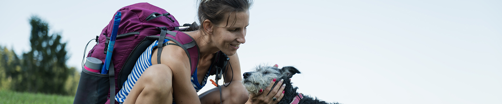 Woman in a blue striped top kneels in a field to pet a small scruffy dog near a purple backpack