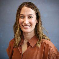 Carleen Acker w Portrait of a smiling woman with shoulder-length light brown hair, wearing an orange blouse, against a blue-gray studio backdrop.