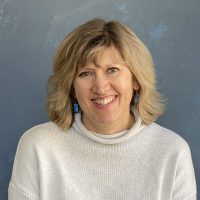 Cathy – Audiologist Smiling blonde woman in a light gray knit sweater, against a textured blue background.