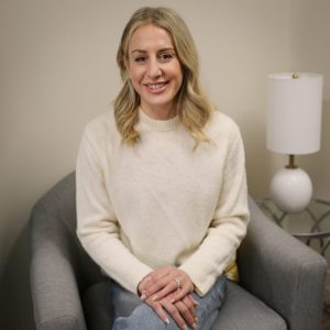 A smiling woman with long blonde hair sits cross‑legged on a gray chair, wearing a cream sweater and jeans, with hands folded in her lap inside a cozy room.